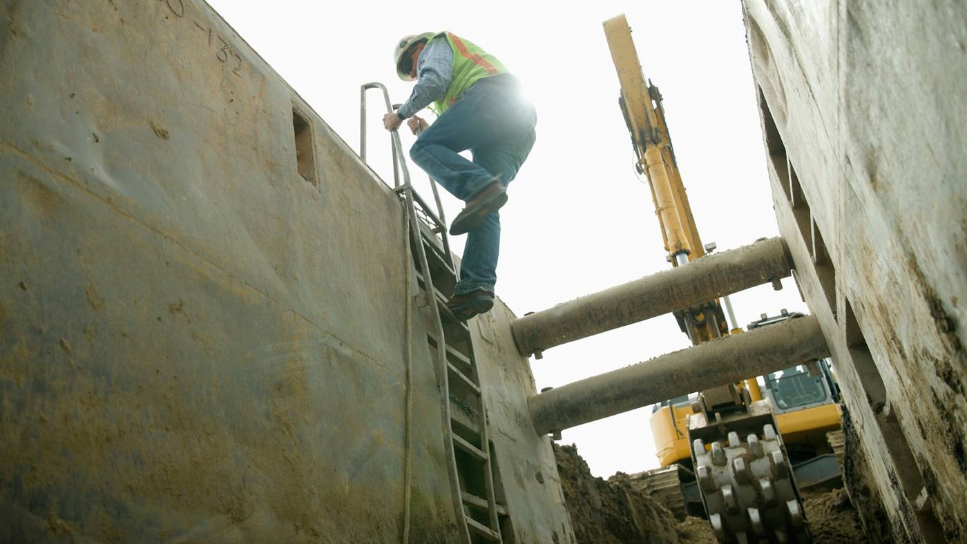A field worker climbing out of a deep utility trench beside an excavator